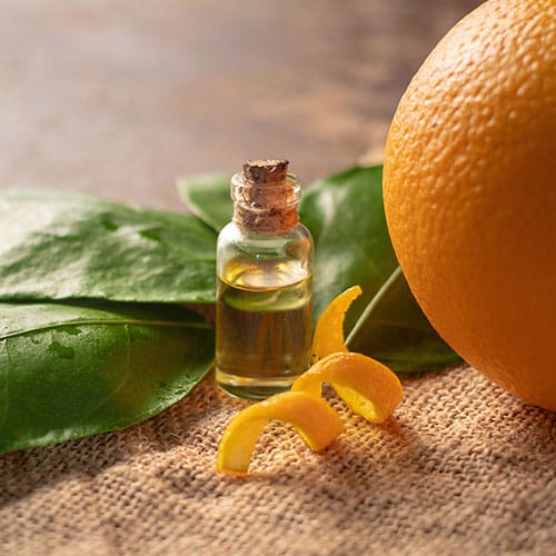 Orangenschalenöl in einem kleinen Glasfläschchen mit Korken. Rechts daneben eine Orange und vor dem Fläschchen ein Stück geschnittene Orangenschale.