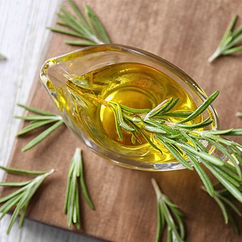 Glasgefäß gefüllt mit Rosmarinöl und einem Zweig frischem Rosmarin auf einem Holzbrett, umgeben von weiteren Rosmarinzweigen.