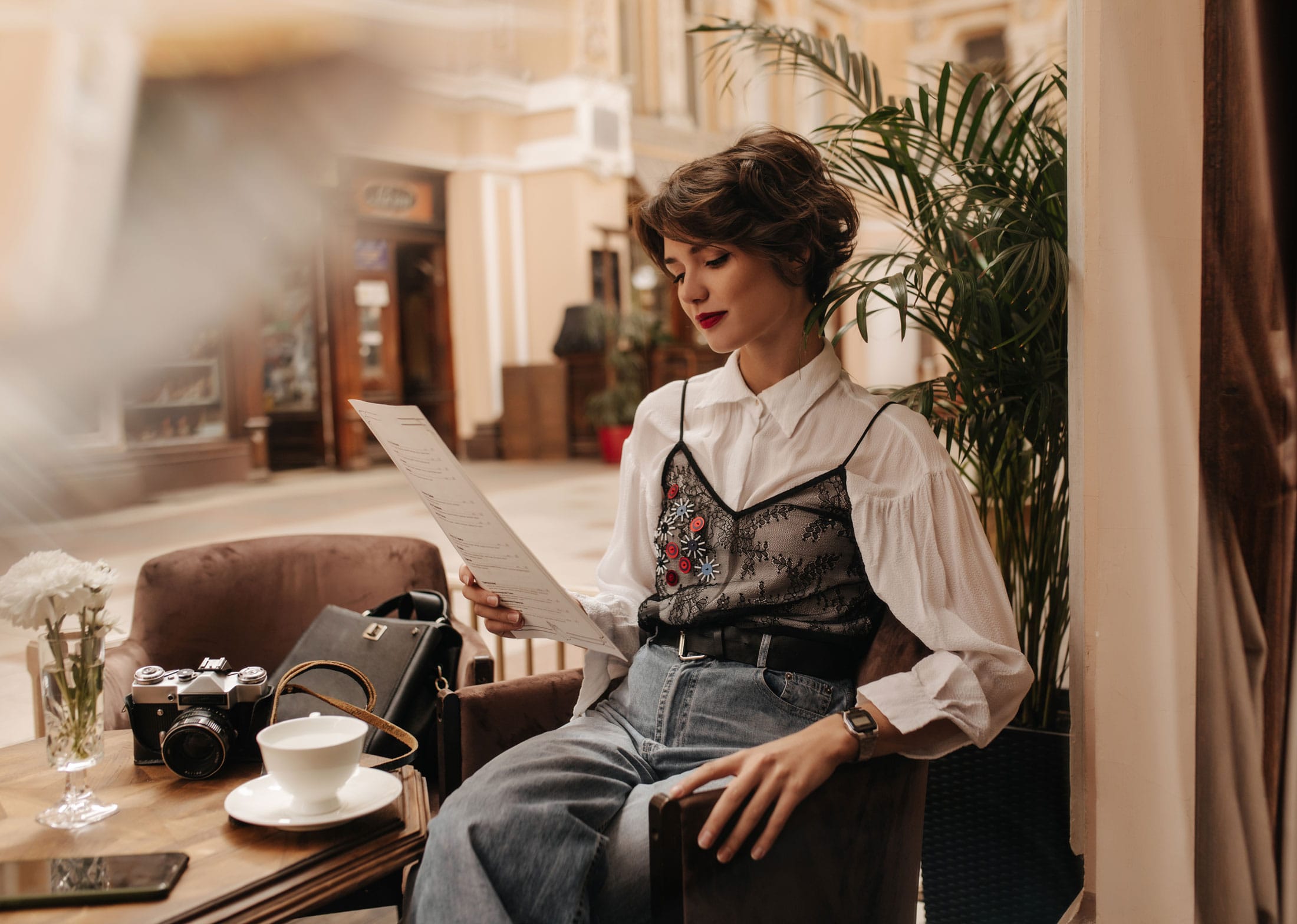 Eine Frau sitzt in einem Café, liest eine Speisekarte und trägt eine weiße Bluse mit besticktem Top und Jeans. Auf dem Tisch stehen eine Kamera, eine Tasse, ein Smartphone und Blumen.