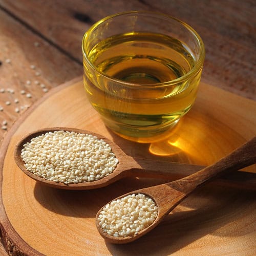 Sesamöl in einer Glasschale, davor zwei Holzlöffel gefüllt mit Sesam